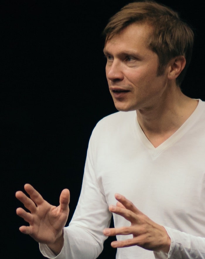 Caspar Schjelbred teaching improvisation, gesturing with open hands while speaking, against a dark studio background.
